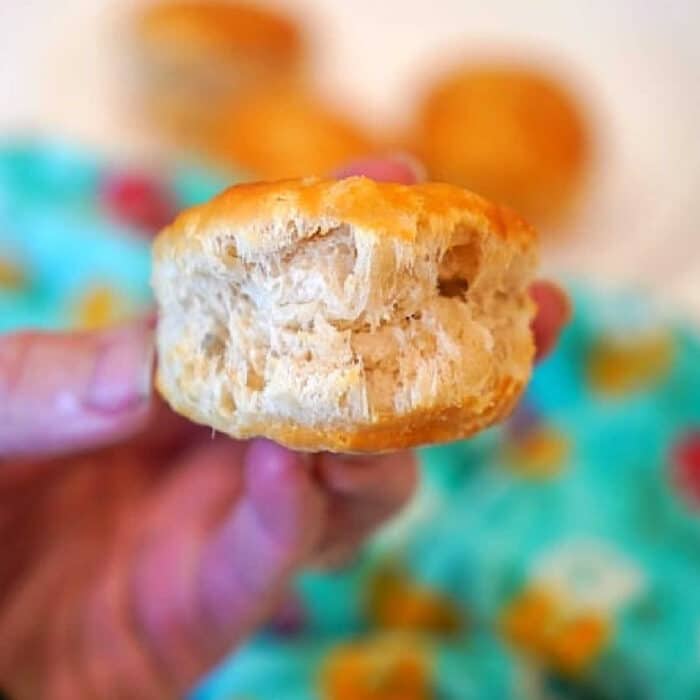 A hand holding a fluffy biscuit with a bite taken out, fresh from frozen biscuits in air fryer, with more biscuits and a turquoise cloth in the background.