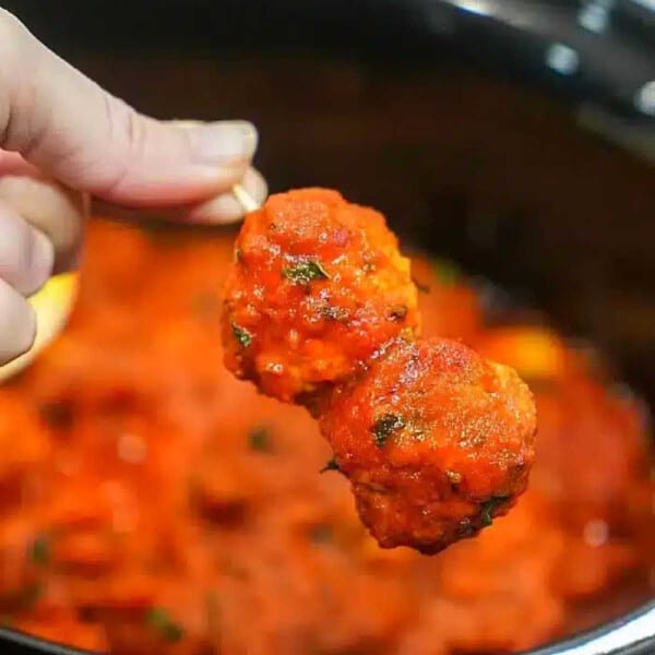 A hand holds two frozen meatballs covered in spaghetti sauce over a pot filled with more sauce, ready to simmer perfectly in the crockpot.