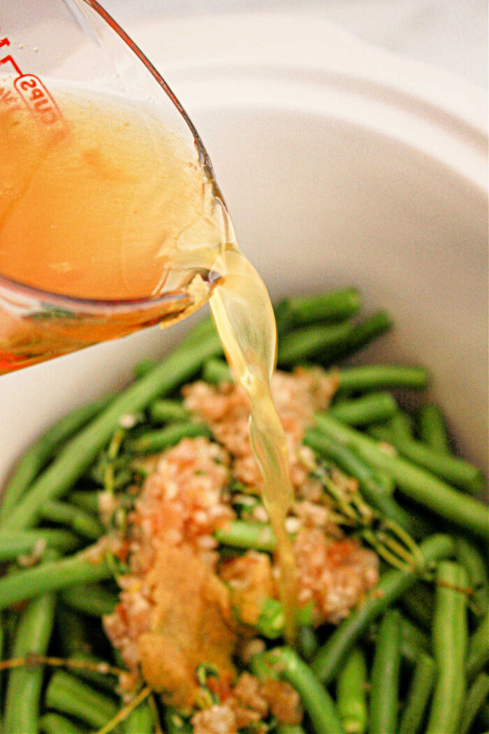 Close-up of a measuring cup pouring broth into a crockpot filled with green beans, minced garlic, and other seasonings. The green beans are uncooked, and the garlic appears to be spread on top.