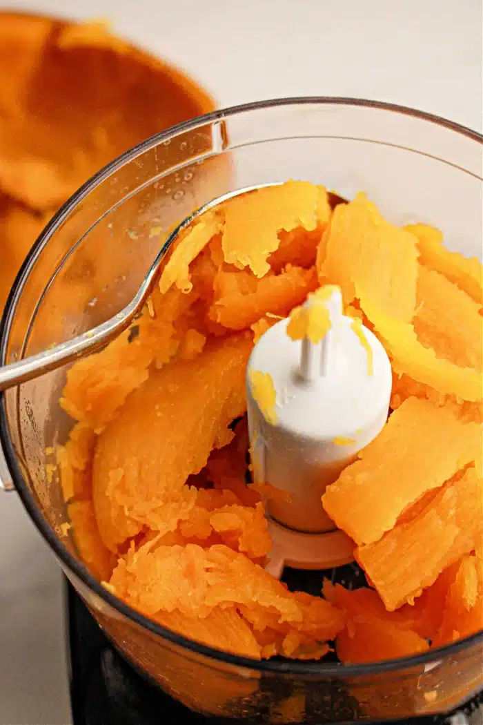 A food processor bowl is filled with chunks of cooked pumpkin. A metal spoon rests on the edge of the bowl. The pieces are bright orange and partially mashed, blending flavors beautifully. The background shows more sweet potato peels, indicating a festive preparation.