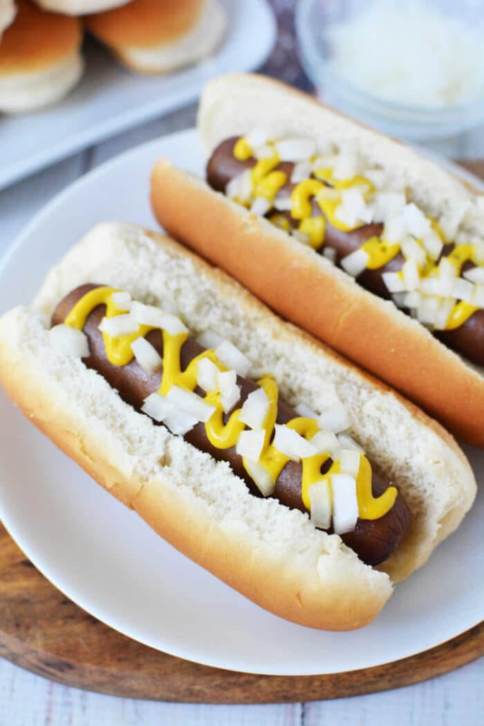 Two hot dogs in buns are placed on a white plate. The hot dogs, slow-cooked to perfection, are topped with diced onions and drizzled with yellow mustard. A platter of additional slow cooker hot dogs and a bowl of chopped onions are visible in the background.