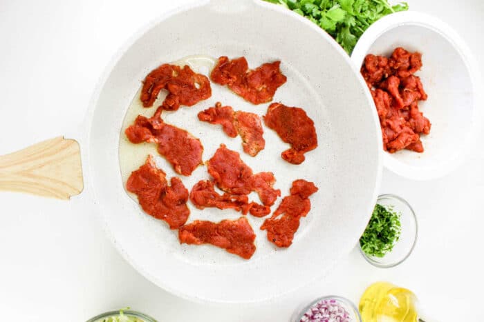 A white frying pan with raw marinated beef slices being cooked. Chopped cilantro, a bowl of additional raw beef, chopped red onions, minced garlic, and a small container of olive oil are arranged around the pan. Some fresh cilantro is also visible, evoking the vibrant setup for Pork Tenderloin Tacos.