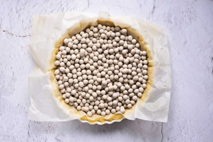 A round pie crust covered with parchment paper is filled with numerous small ceramic baking beads, preparing the foundation for a savory sweet potato pie with sweetened condensed milk. The pie is placed on a white, slightly textured surface.