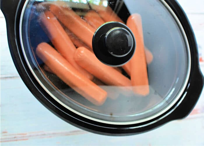 A black slow cooker with several slow cooker hot dogs inside, viewed from the top. The glass lid partially obscures the hot dogs. The slow cooker rests on a light-colored surface.