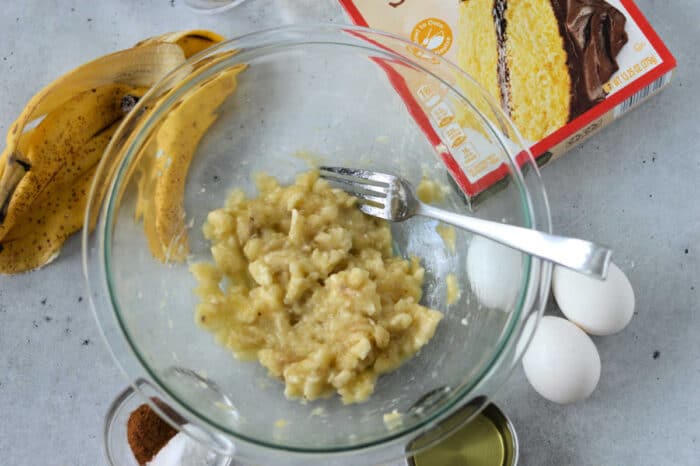 A mixing bowl with mashed bananas and a fork sits on a countertop. Surrounding the bowl are two eggs, a box of cake mix, banana peels, a small bowl of cinnamon, and a can of baking ingredients—all the essentials for an easy banana cake recipe with cake mix.