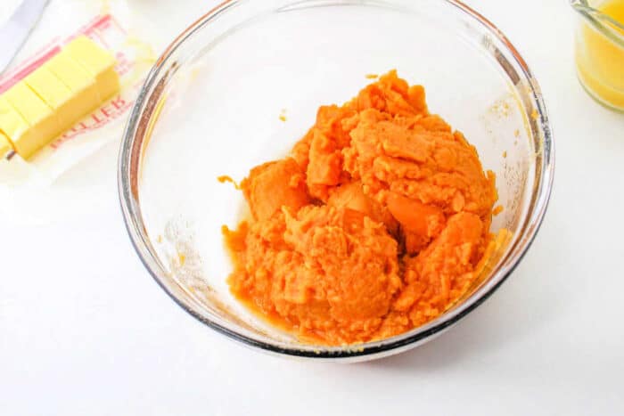 A glass bowl filled with mashed sweet potatoes is on a white surface. Next to it, there is a package of partially sliced butter and a small jar containing a yellow liquid, perfect ingredients for your recipe for Sweet Potato Pie with Sweetened Condensed Milk.