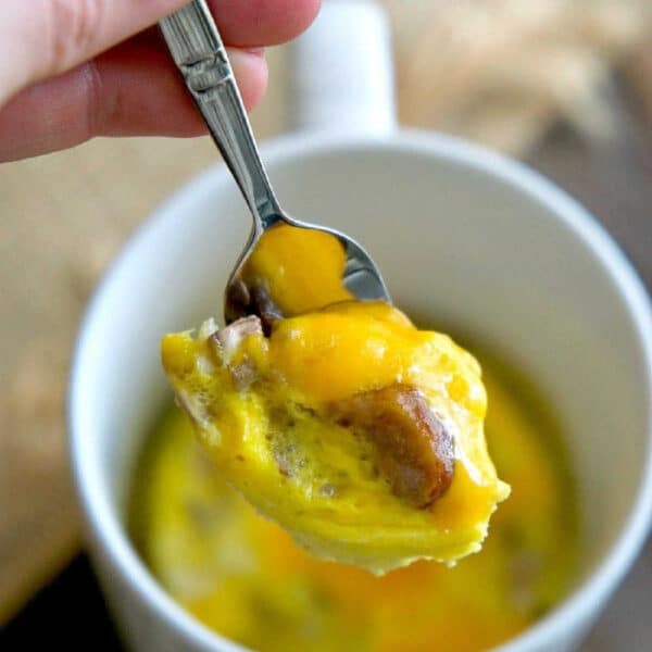A hand holds a spoon with cheesy egg and sausage above a white mug filled with omelette in a mug.