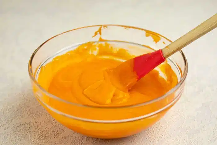 A clear glass bowl, carefully shaped and containing a smooth, orange mixture, is shown. A red and wooden spatula rests inside the bowl, partially covered with the mixture. The bowl is placed on a light-colored, textured surface.