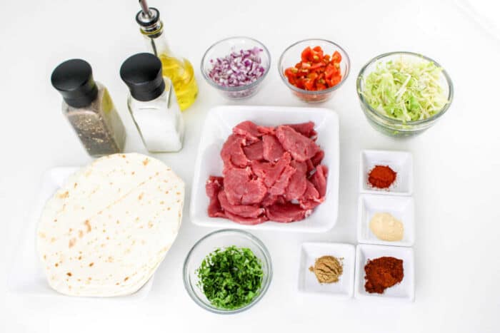 Ingredients for a meal are arranged on a white surface, including tortillas, raw beef slices, chopped cilantro, shredded cabbage, diced red onions, chopped red peppers, various spices in small dishes for making Pork Tenderloin Tacos, a bottle of olive oil, and salt and pepper shakers.