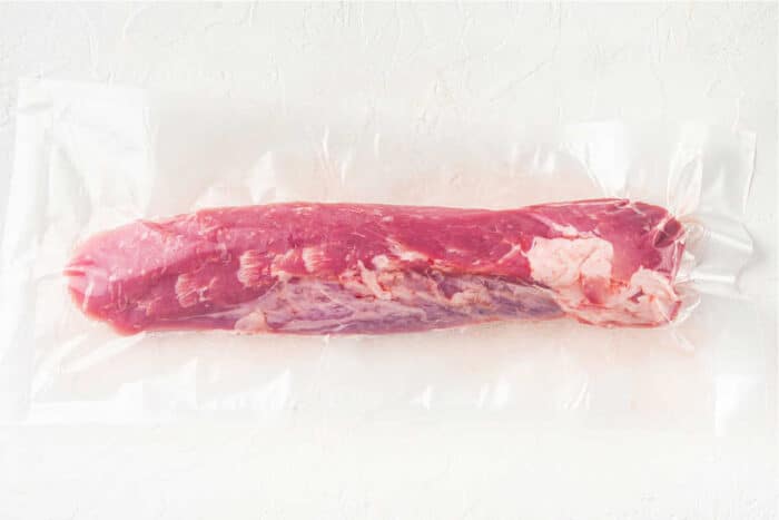 A vacuum-sealed package containing a raw, whole pork tenderloin on a plain, light-colored surface. The meat is pink with visible marbling and fat along its side—perfect for making mouthwatering Pork Tenderloin Tacos.