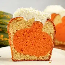 A single piece of cake on a white plate reveals an orange pumpkin shape inside the cake. Topped with a layer of white frosting, the cross-section showcases the charming design. In the blurred background, another piece of cake awaits its turn.