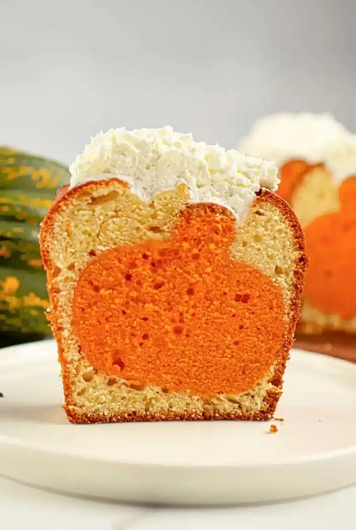 A single piece of cake on a white plate reveals an orange pumpkin shape inside the cake. Topped with a layer of white frosting, the cross-section showcases the charming design. In the blurred background, another piece of cake awaits its turn.