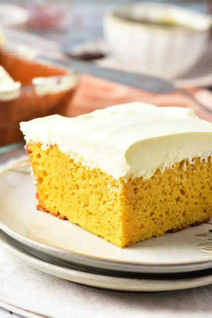 A close-up image of a slice of yellow pumpkin cake with a thick layer of white frosting on a plate. The background includes a blurred view of more frosted cake and tableware, including a knife and a bowl. The scene is set on a light-colored tablecloth.