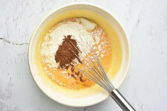 A mixing bowl contains a mixture of yellow liquid, likely beaten eggs, and a heap of dry ingredients, including flour and cocoa powder. A silver whisk is resting inside the bowl, positioned toward the right side. It looks like the beginnings of a pumpkin cake with yellow cake mix on a light-colored surface.