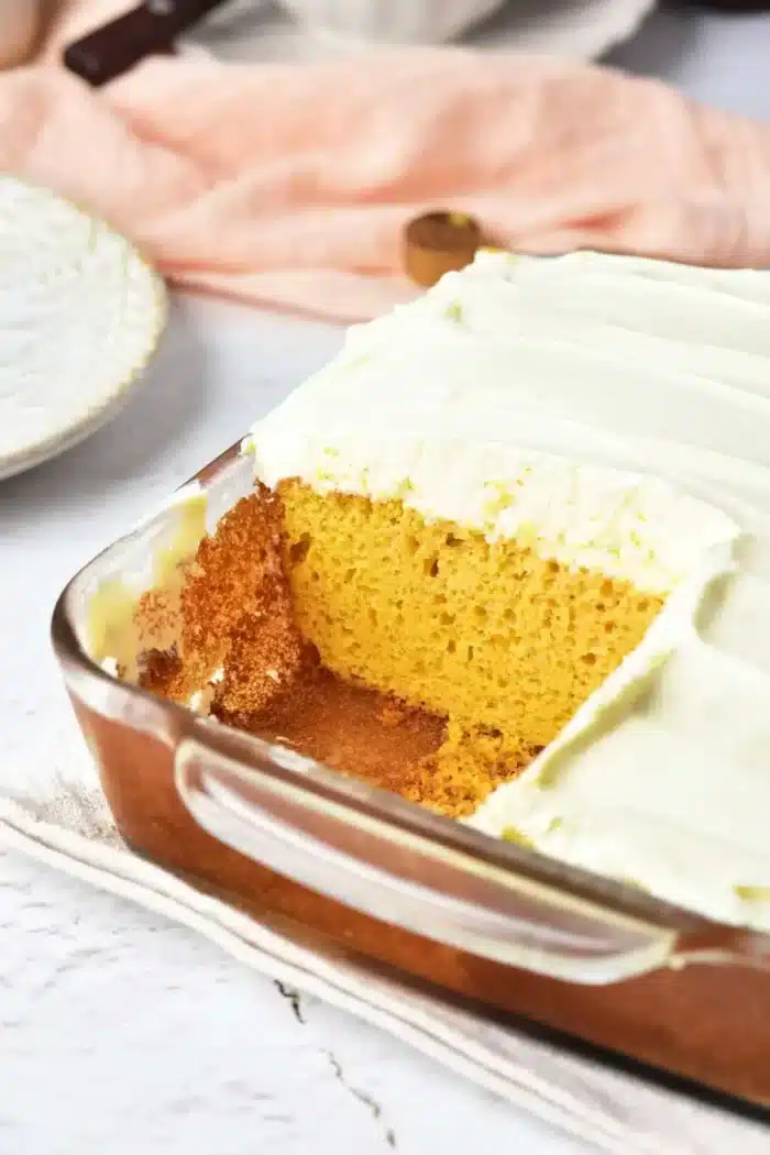 A rectangular glass baking dish contains a pumpkin cake with yellow cake mix, topped with white frosting. A square piece has been removed, revealing the cake's interior. The dish sits on a light-colored cloth, with a plate and peach-colored fabric in the background.