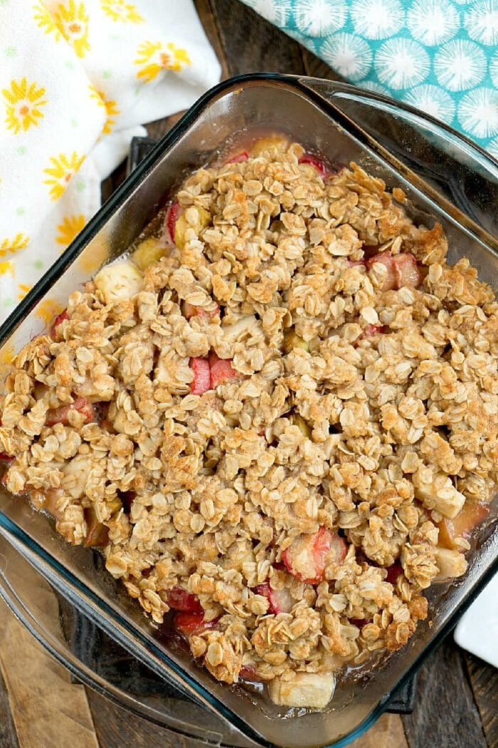 This apple rhubarb crunch dessert is amazing! Using fresh or frozen fruit with an oatmeal crisp topping it's great for brunch, breakfast or a light dessert. #rhubarb #crisp #desserts #dessert #crisp #crunch #apple #oatmeal A glass dish filled with baked rhubarb crunch, topped with golden oat streusel, sits on a wooden table.