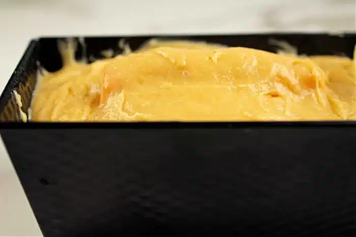 A close-up of thick yellow batter poured into a rectangular black baking pan. The batter appears smooth and fills the pan almost to the top, ready for baking, hinting at the delicious shape inside the cake to come.