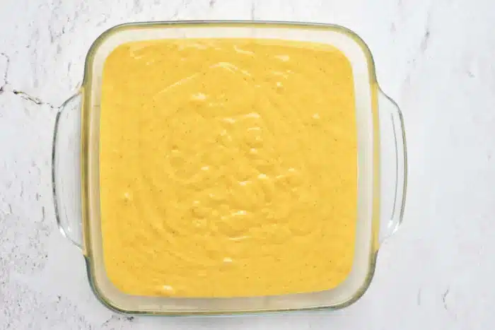 A square glass dish filled with a smooth, yellow batter sits on a light-colored marbled surface. The batter, perfect for pumpkin cake with yellow cake mix, has a few small bubbles scattered across the top, indicating that it has just been prepared and is ready to be baked.