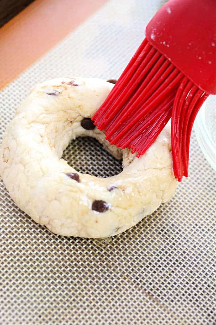 A red pastry brush applies liquid to an uncooked chocolate chip 2 Ingredient Bagel on a baking mat.