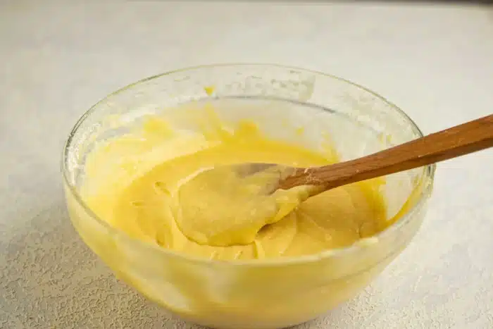 A glass bowl filled with yellow cake batter holds a wooden spoon with some batter on it, hinting at the shape inside the soon-to-be baked cake. The background features a light-colored countertop.