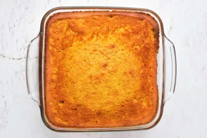 A square glass baking dish containing a freshly baked, golden-brown pumpkin cake with yellow cake mix sits on a light, textured surface. The edges of the cake appear slightly darker and crisp. The pumpkin cake has a smooth, even surface.