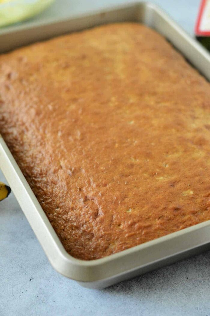 A rectangular banana cake, baked to a golden color using a cake mix, is shown in a metal baking pan. The cake has a smooth, slightly textured surface, and the edges are slightly raised. The background is a light-colored countertop.