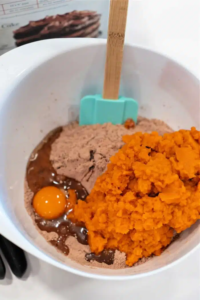 A white mixing bowl contains ingredients for pumpkin brownies. Visible items include a raw egg, vibrant pumpkin puree, and rich brown cake mix. A blue spatula and a wooden one are also in the bowl, with a recipe card slightly visible in the background.