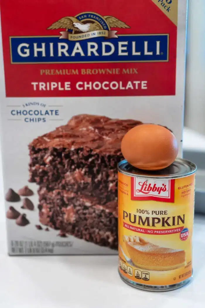 A box of Ghirardelli triple chocolate brownie mix is cleverly paired with a can of Libby's 100% pure pumpkin, hinting at the delightful creation of pumpkin brownies. An egg perches atop the can, ready to bring this delicious idea to life.