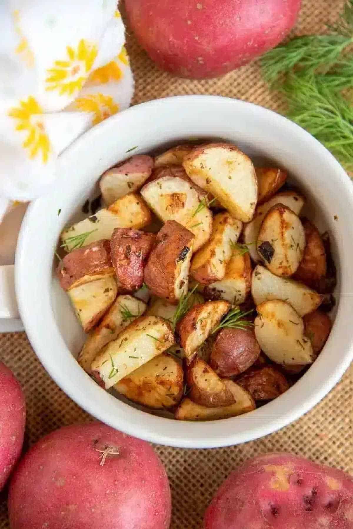 Air fryer red potatoes with dill and garlic are the best side dish with any dinner. Inexpensive yet full of flavor, you've got to try these tonight. A white bowl filled with air fryer red potatoes, garnished with herbs, surrounded by whole red potatoes.
