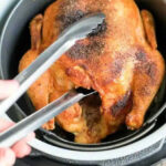 An air fryer whole chicken sits in the basket, perfectly cooked and golden, as metal tongs lift it up for serving.