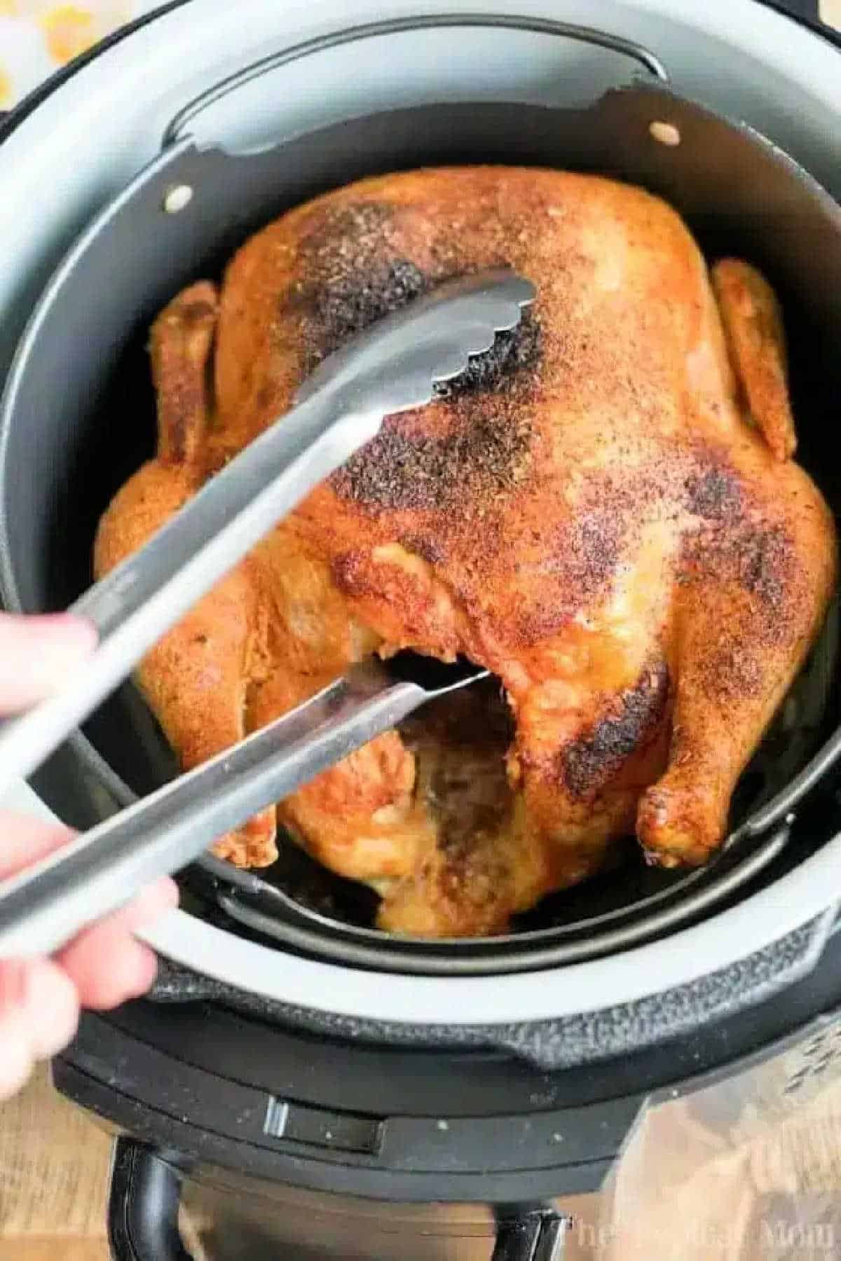 Air fryer whole chicken is amazing! Super crispy skin outside and tender meat inside makes the best dinner ever. Give this easy air fryer recipe a try. #airfryerrecipes #airfryer #chicken #wholechicken #crispy #thetypicalmom #ninjafoodirecipes #ninjafoodi An air fryer whole chicken sits in the basket, perfectly cooked and golden, as metal tongs lift it up for serving.