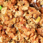 Close-up of a dish featuring baked stuffing with pieces of bread, visible chunks of onion, and herbs—perfect for those wondering how to cook stuffing in the oven. A metal serving spoon is partially visible, and a sprig of parsley is seen in the corner. The texture appears crispy and moist.