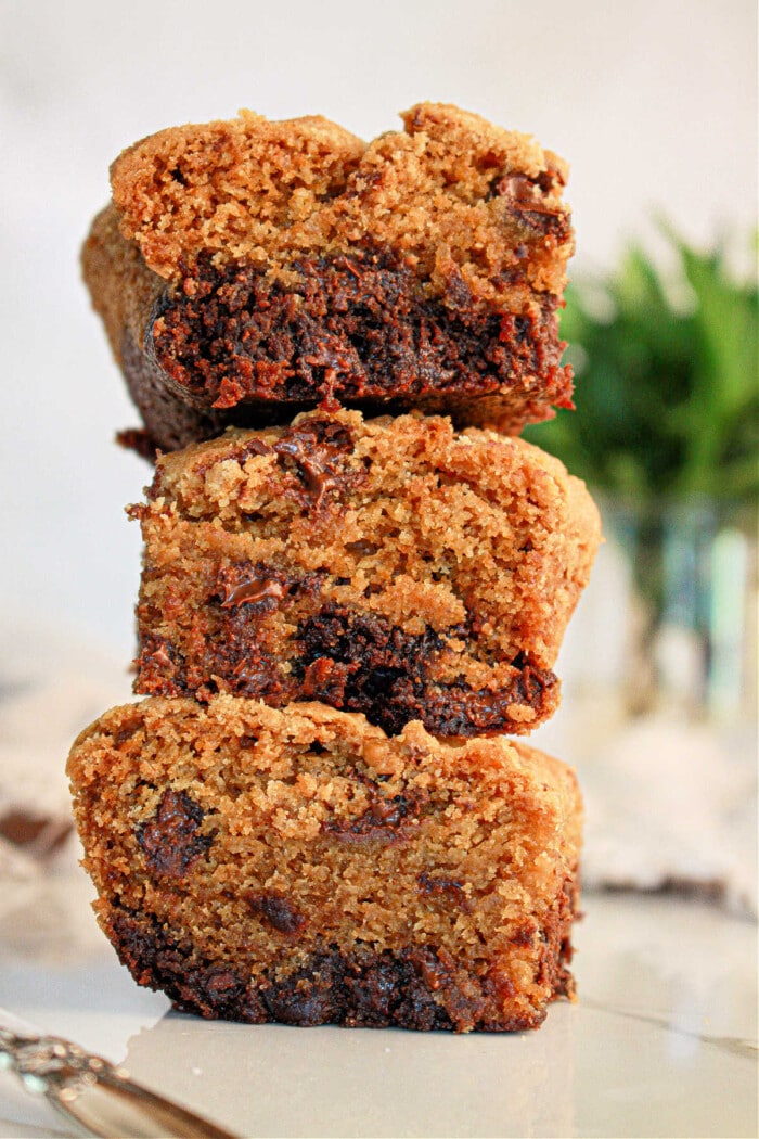 Three stacked slices of brookies, rest on a white surface. The bread appears moist and fluffy, with visible chocolate chunks. A blurred green plant in the background adds a touch of color to the scene.