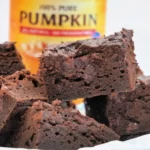 Close-up of 3 ingredient pumpkin brownies stacked in front of a can of pure pumpkin puree. The moist and fudgy treats contrast beautifully with the yellow can's red label in the backdrop.