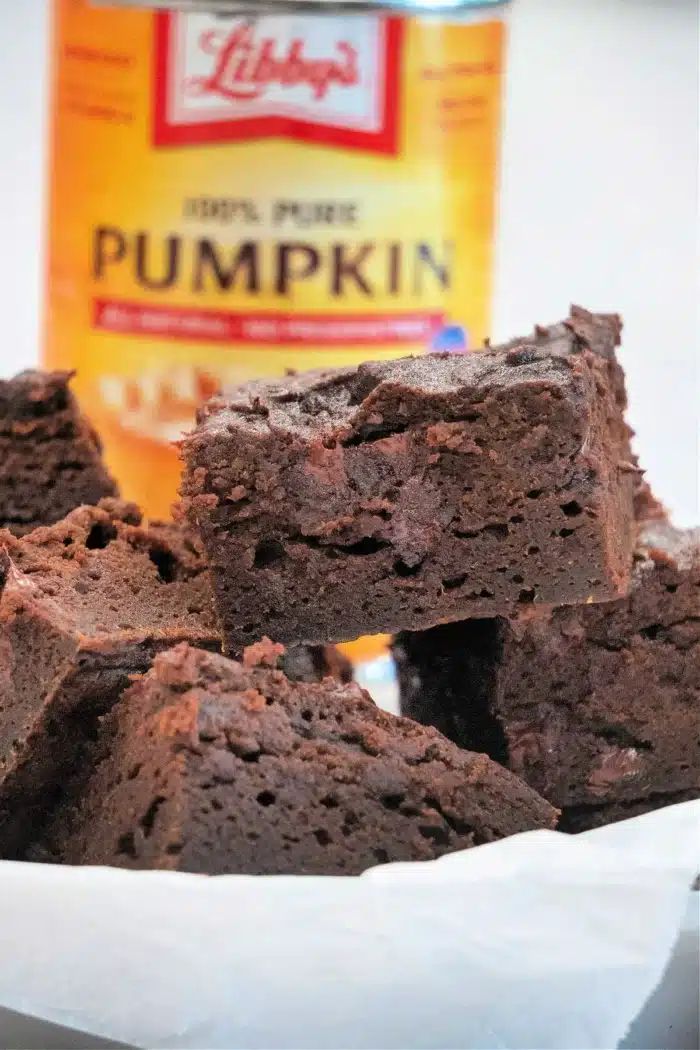 A close-up of decadent chocolate brownies stacked on a white plate, showcasing their rich texture. In the background, a can labeled "Libby's 100% Pure Pumpkin" adds a hint of autumn charm to this dessert scene.