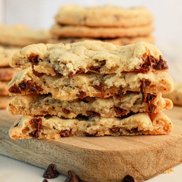 A stack of chocolate chip cookies made with pancake mix is halved and placed on a wooden board. The cookies are thick with visible chunks of melted chocolate. Some chocolate chips are scattered around the board, and more pancake mix cookies are stacked in the background.