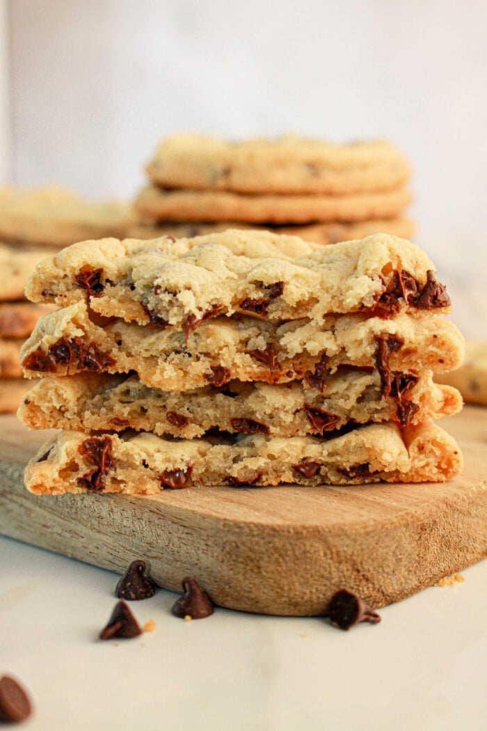 A stack of chocolate chip cookies made with pancake mix is halved and placed on a wooden board. The cookies are thick with visible chunks of melted chocolate. Some chocolate chips are scattered around the board, and more pancake mix cookies are stacked in the background.