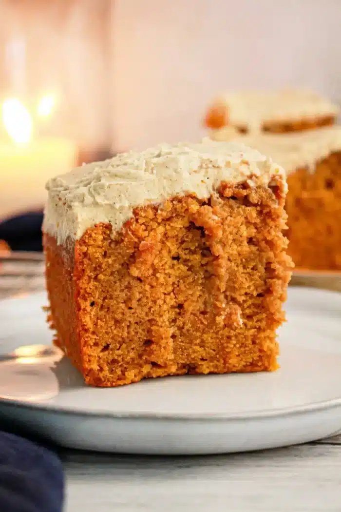 A close-up of a slice of Pumpkin Poke Cake with cream cheese frosting on a white plate. The cake has a golden-brown color and a moist texture, while the thick frosting covers the top of the slice. A blurred background shows another slice and a lit candle.
