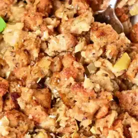 Close-up of a dish of oven-cooked stuffing, showcasing browned bread cubes mixed with cooked onions and herbs. The texture appears moist and chunky, with a spoon partially visible in the top right corner. Garnished with a small parsley leaf.