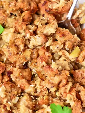 Close-up of a dish of oven-cooked stuffing, showcasing browned bread cubes mixed with cooked onions and herbs. The texture appears moist and chunky, with a spoon partially visible in the top right corner. Garnished with a small parsley leaf.