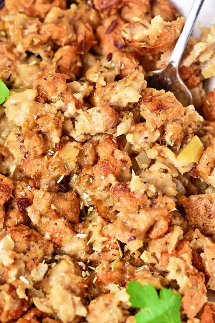 Close-up of a dish of oven-cooked stuffing, showcasing browned bread cubes mixed with cooked onions and herbs. The texture appears moist and chunky, with a spoon partially visible in the top right corner. Garnished with a small parsley leaf.