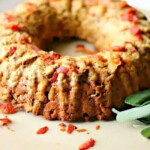 A savory bundt cake with herbs and bacon pieces on a beige plate, garnished with fresh sage leaves, showcases how to cook stuffing in a bundt pan, elevating this classic dish to new heights.