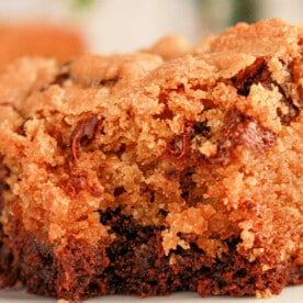 Close-up of a moist slice of chocolate chip cookie cake on a white plate, resembling the perfect blend achieved when learning how to make brookies with brownie mix and cookie dough. The golden-brown crust and melted chocolate chips create a deliciously textured and rich appearance.