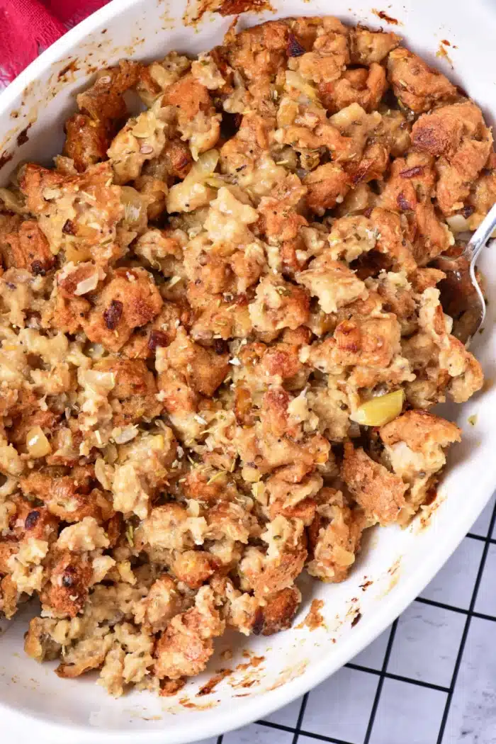 A close-up of a casserole dish filled with baked stuffing showcases the art of how to cook stuffing in the oven. The golden brown top reveals pieces of bread, herbs, and bits of sautéed onions and celery. A silver serving spoon is partially visible on the right side, ready to serve.