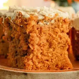 A slice of moist orange-brown Pumpkin Poke Cake with a light-colored frosting sits on a wooden plate. The cake's texture appears crumbly and dense. In the background, there are orange-hued flowers slightly out of focus, enhancing the warm, cozy environment.