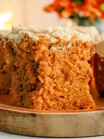 A slice of moist orange-brown Pumpkin Poke Cake with a light-colored frosting sits on a wooden plate. The cake's texture appears crumbly and dense. In the background, there are orange-hued flowers slightly out of focus, enhancing the warm, cozy environment.