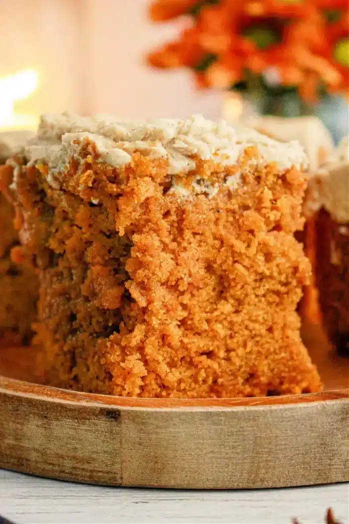 A slice of moist orange-brown Pumpkin Poke Cake with a light-colored frosting sits on a wooden plate. The cake's texture appears crumbly and dense. In the background, there are orange-hued flowers slightly out of focus, enhancing the warm, cozy environment.