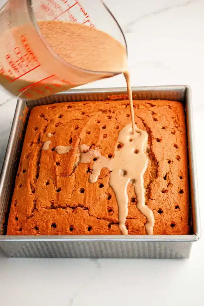 A square Pumpkin Poke Cake in a metal baking pan is being drizzled with a light brown liquid from a measuring cup. The cake has multiple holes poked into its surface to allow the liquid to seep in, ensuring every bite is moist and flavorful.