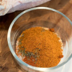 A glass bowl filled with a mix of dry spices for a smoked turkey rub recipe sits on a wooden surface next to raw poultry.