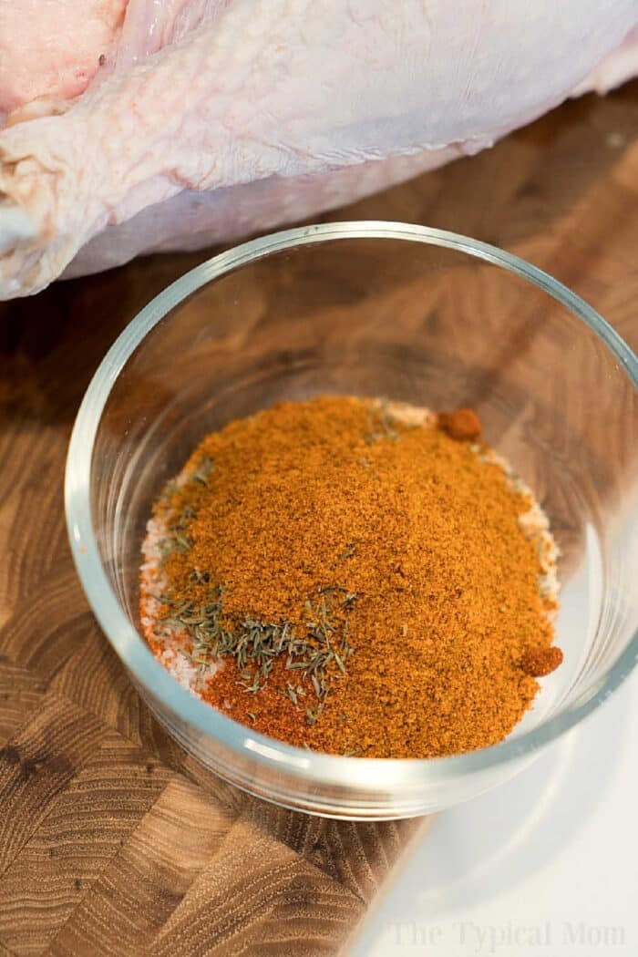 A glass bowl filled with a mix of dry spices for a smoked turkey rub recipe sits on a wooden surface next to raw poultry.